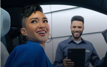 Woman smiling from inside a car while a man outside holds a tablet, appearing to assist or provide customer service.
